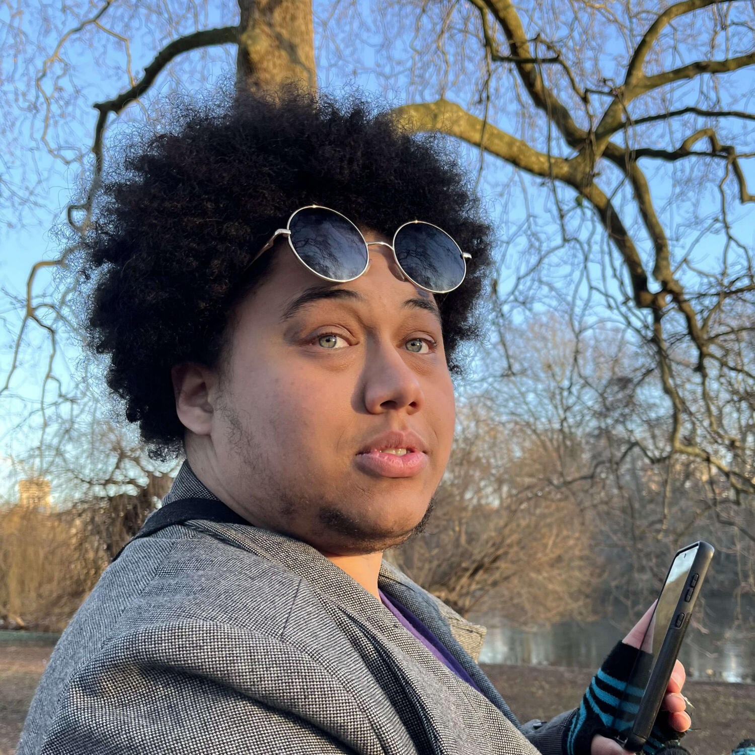 a photograph of Luc, looking offscreen. he is wearing a grey coat over a purple jumper, with black and blue fingerless gloves and black sunglasses on his forehead.